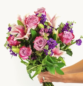 A vibrant bouquet featuring pink lilies, purple flowers, and green foliage, elegantly held.