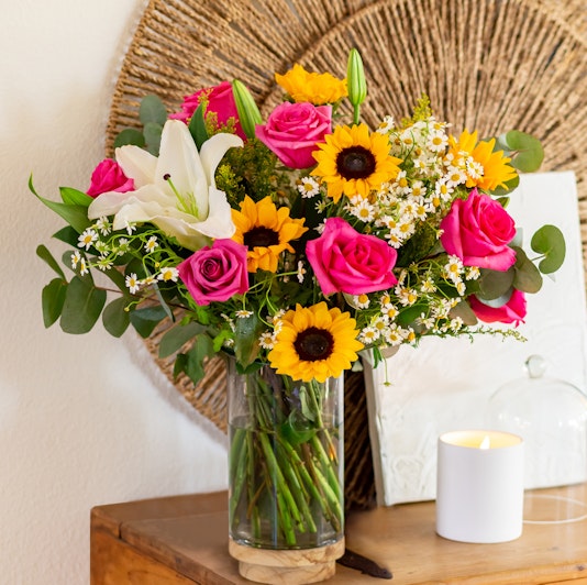 Vibrant floral arrangement featuring pink roses, sunflowers, and white lilies in a glass vase.