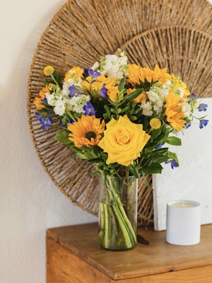 Bright bouquet of yellow roses, sunflowers, and blue flowers in a stylish glass vase.