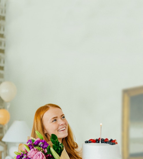 A joyful woman celebrating with a birthday cake and colorful flowers in a cozy setting.
