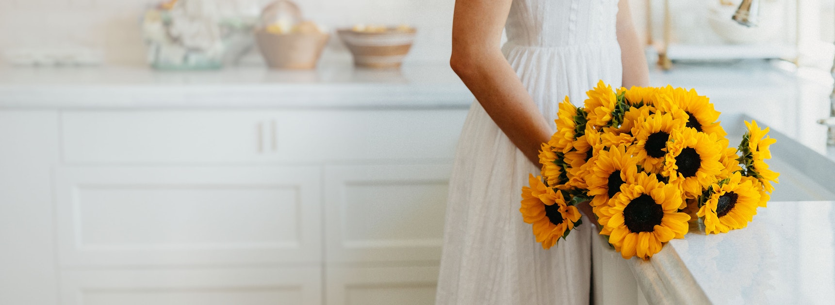 A cheerful bouquet of vibrant sunflowers held by a woman in a white dress, brightening a kitchen.