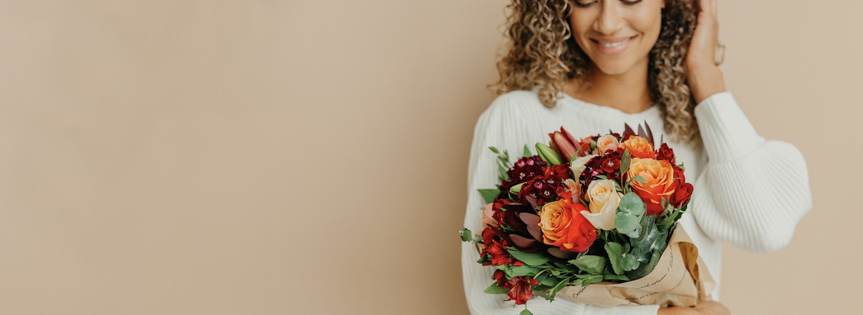 Smiling woman in a cozy sweater holding a vibrant mixed flower bouquet against a neutral backdrop.