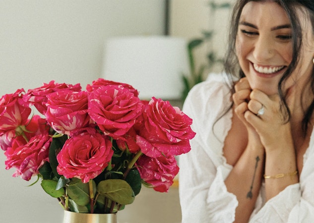 A vibrant bouquet of pink roses sits on a table as a woman smiles joyfully nearby.