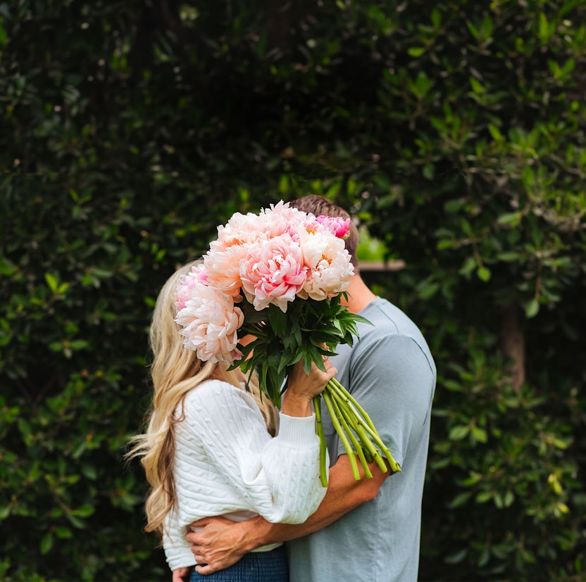 A couple standing together holding a vibrant bouquet of peonies, surrounded by greenery.