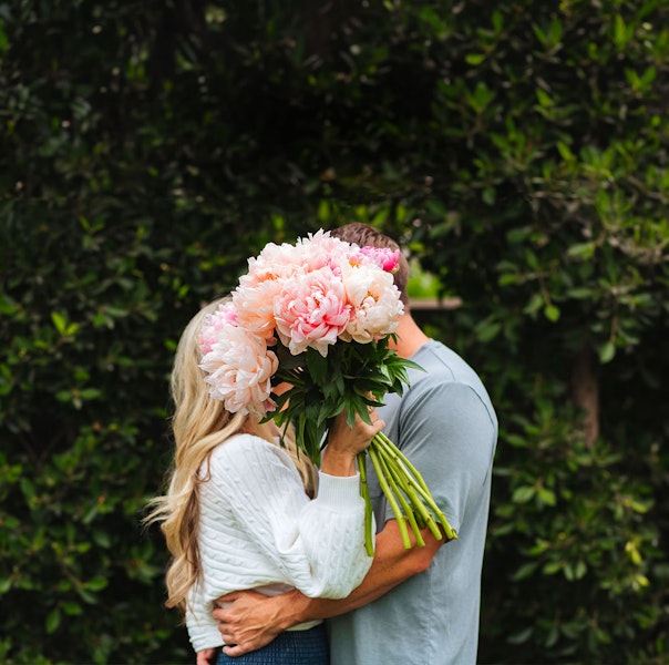 A couple standing together holding a vibrant bouquet of peonies, surrounded by greenery.
