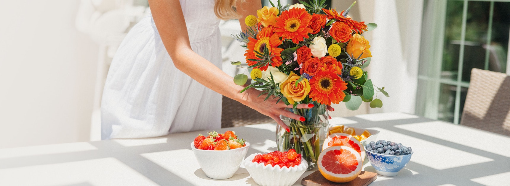 Vibrant floral arrangement with orange gerbera daisies and roses, complemented by fresh fruits.